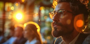 Bearded man with glasses looking thoughtful, bathed in warm sunlight, with blurred people in the background.