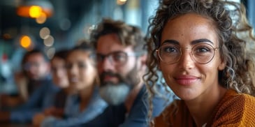 Smiling woman with curly hair and glasses, wearing a mustard sweater, sitting with a group of people in a warmly lit indoor setting.