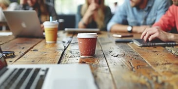 Coffee cups, laptops, and notebooks on a rustic wooden table with people blurred in the background, suggesting a casual meeting or workspace.